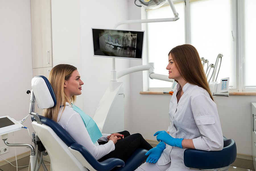 Woman wearing a white top sitting in an exam chair discussing dental treatment options with her female dentist. 